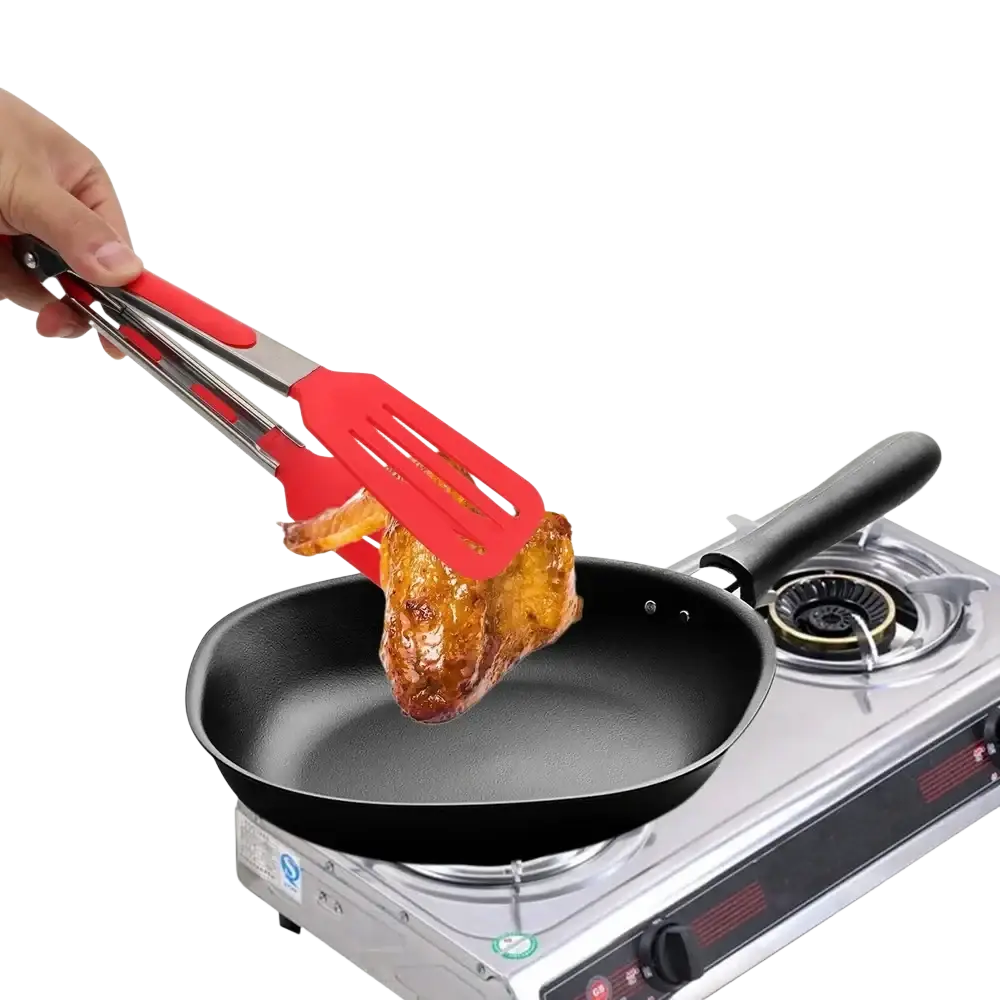 Red silicone kitchen tongs with stainless steel handles lifting roasted chicken from a black non-stick frying pan