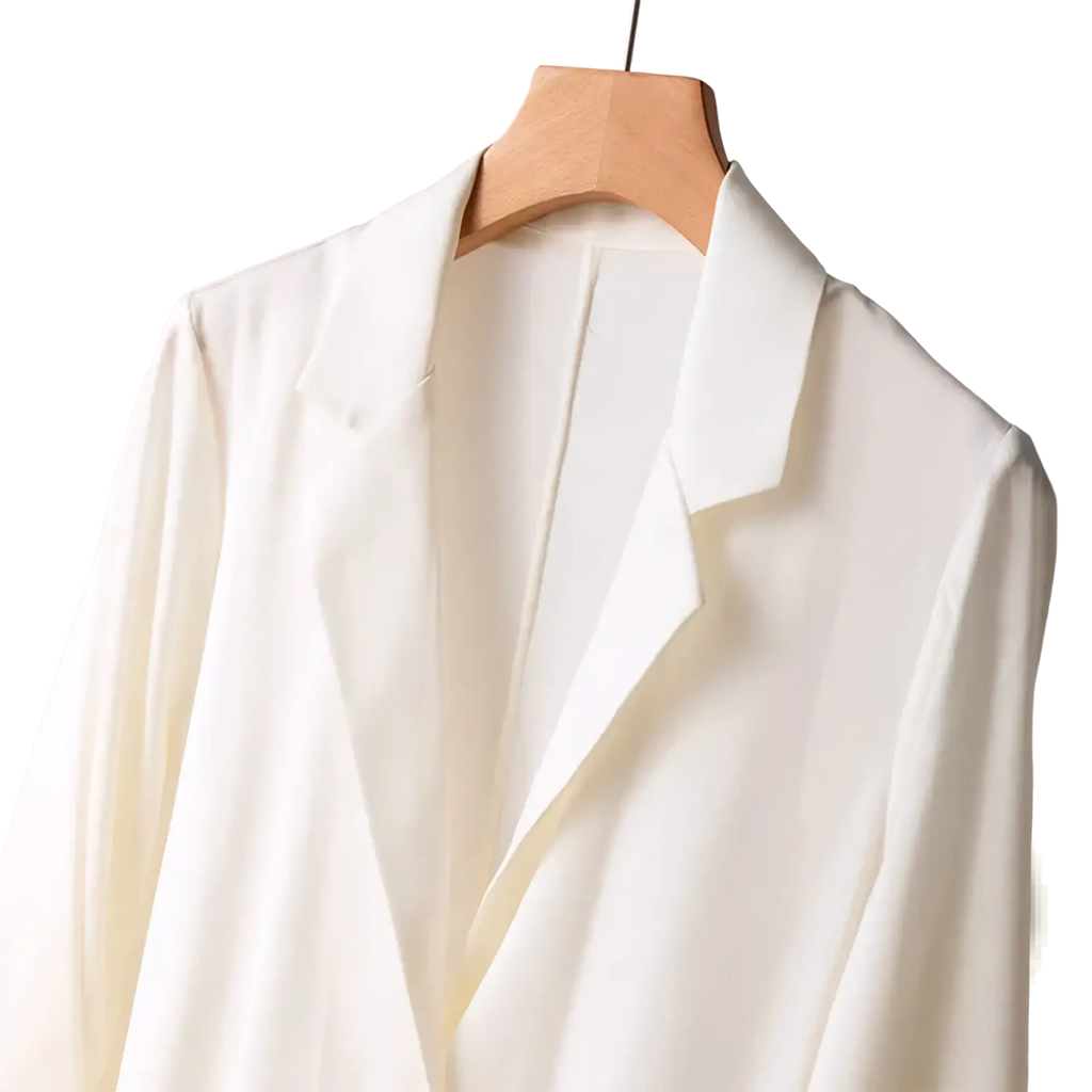 A sleek off-white silk blazer with smooth satin finish and one button hanging elegantly on a wooden hanger