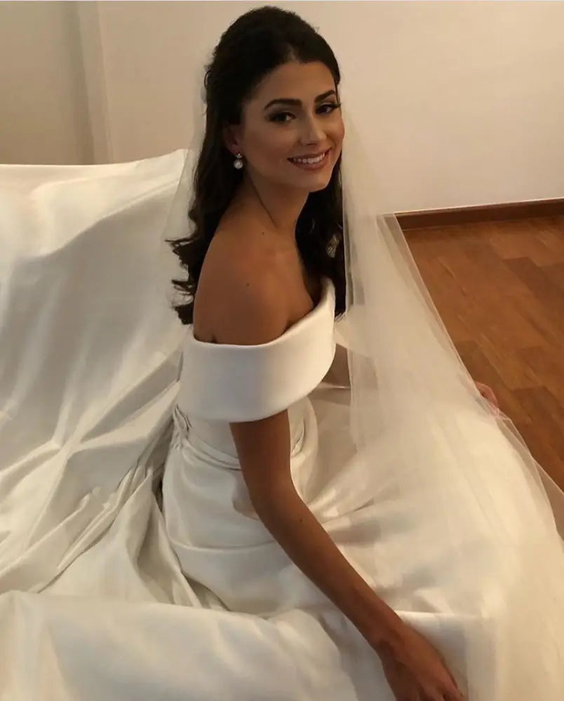 A flowing white satin off the shoulder a line wedding gown with a fitted bodice and sweep train
