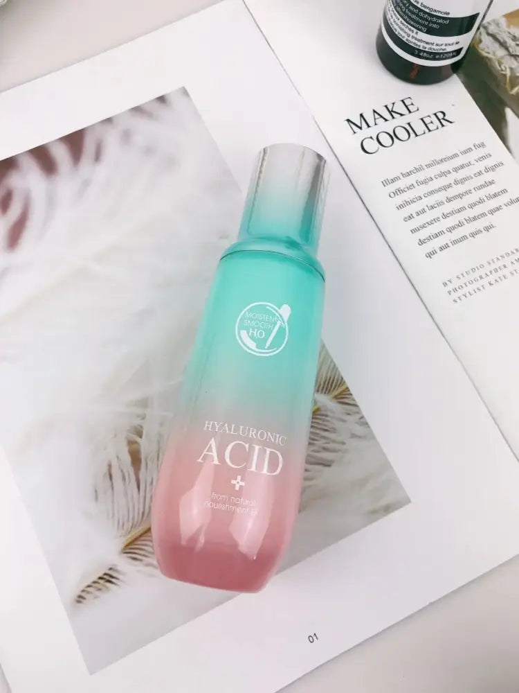 A gradient-tinted glass bottle of hyaluronic acid serum with a metallic silver cap in a hydrating skincare gift set
