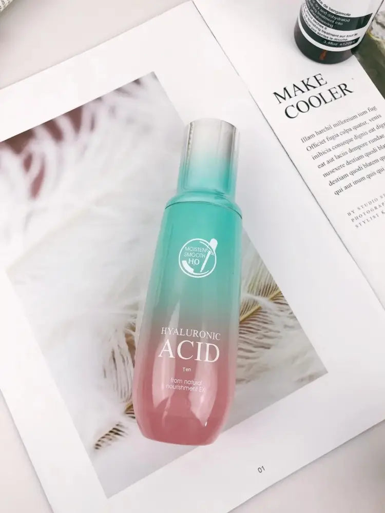 A gradient teal to pink glass bottle of hyaluronic acid serum with a silver cap in a hydrating skincare gift set