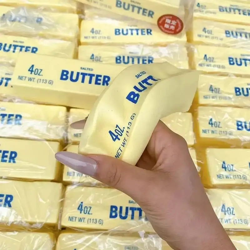 A hand holds a pale yellow 4oz salted butter stick stress relief toy with blue printed labeling and softened surface
