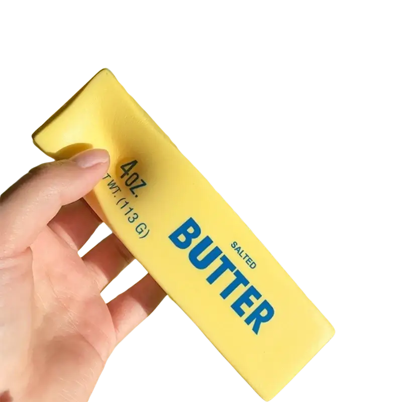 A hand holds a yellow salted butter stick stress relief toy with blue lettering labeled 4oz 113g in bold font
