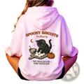 A soft pink hoodie featuring a halloween graphic of a black cat kneading dough with spooky biscuits bakery text