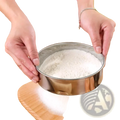 Circular stainless steel flour sifter with hammered texture sifting white flour onto wooden board for baking use