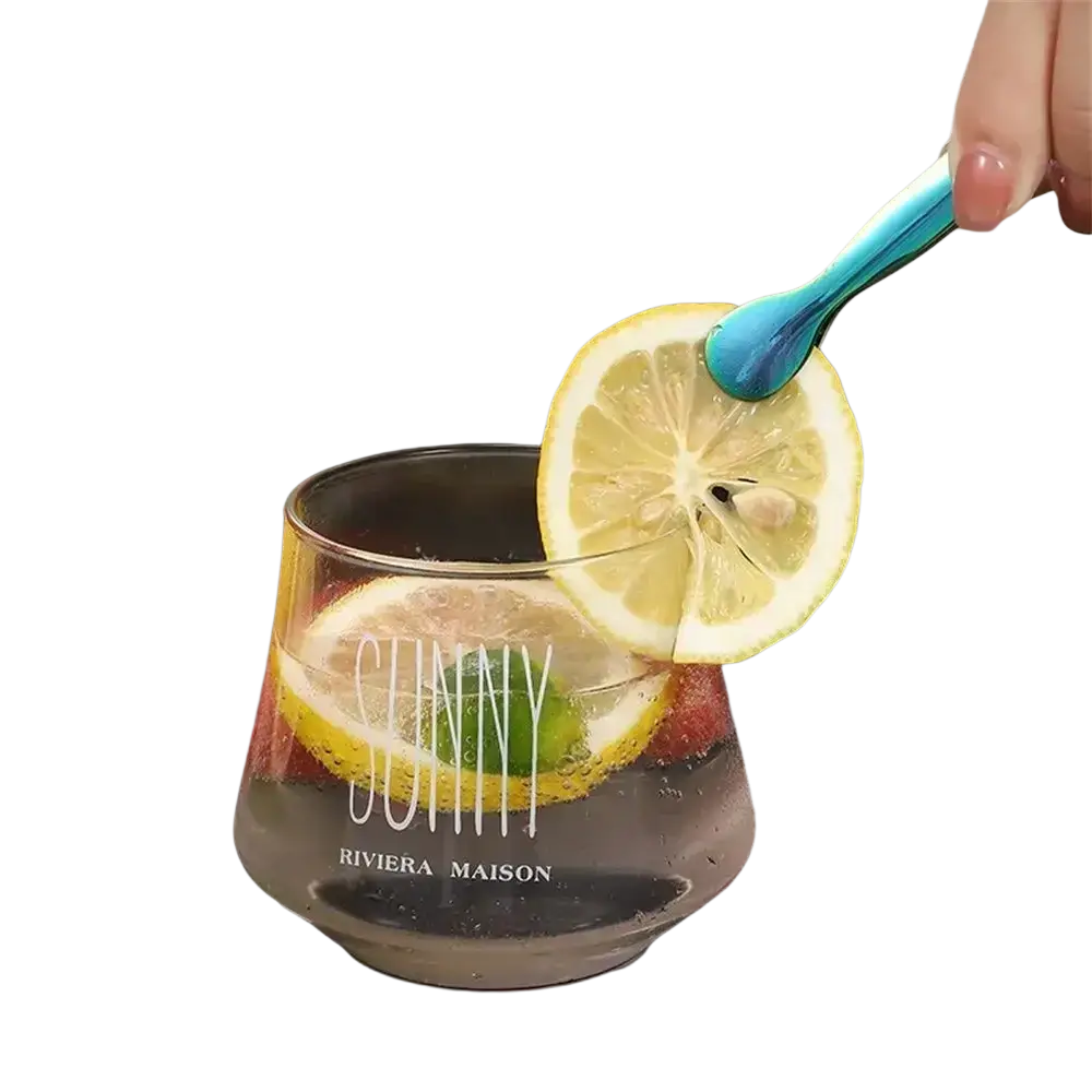 A clear glass tumbler with sunny and riviera maison print filled with a sparkling beverage and lemon slices next to stainless steel mini ice tongs for kitchen and bar