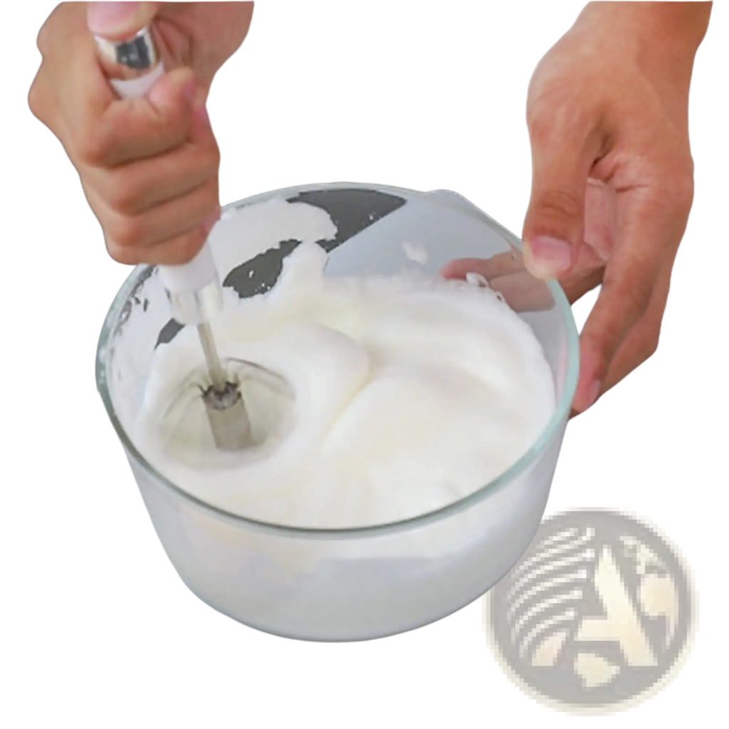 A clear glass bowl with whipped egg whites stirred by a stainless steel semi automatic rotating egg beater whisk