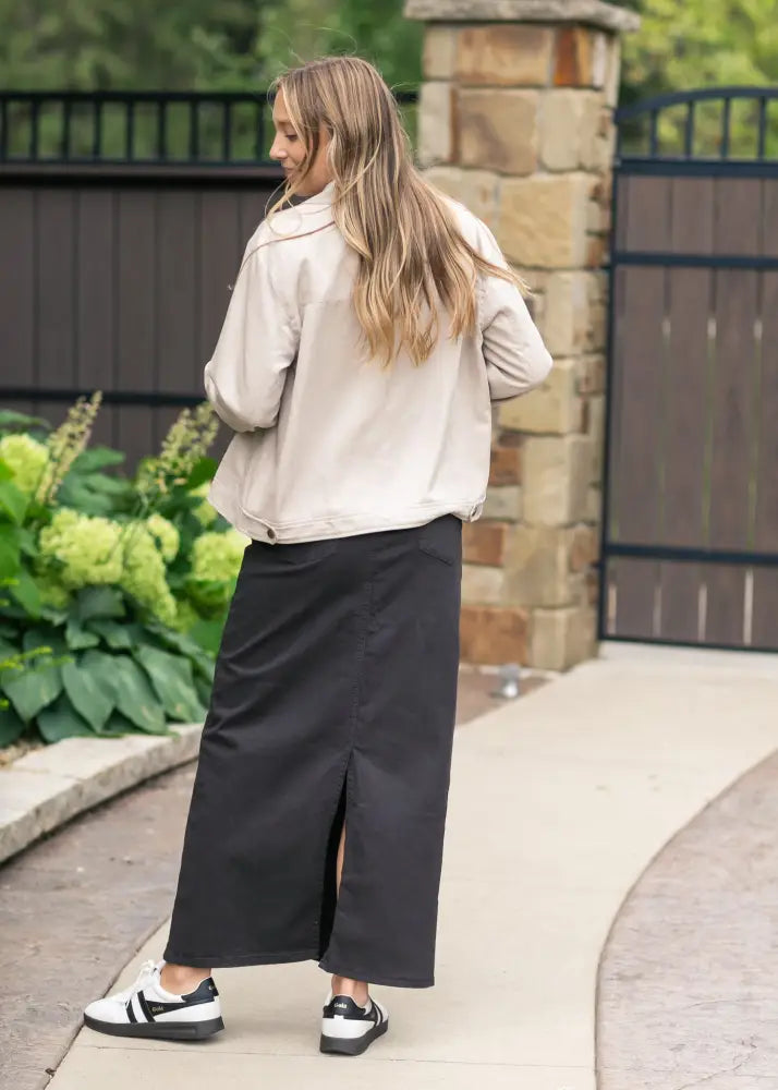 A woman wearing a beige jacket and black wide-leg trousers with a front slit paired with sneakers in a Stella Onyx black high waisted denim maxi skirt product