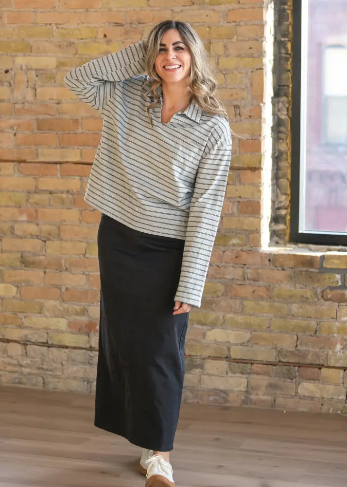 A woman wears a long-sleeved striped polo shirt with navy lines paired with a Stella Onyx black high waisted denim maxi skirt
