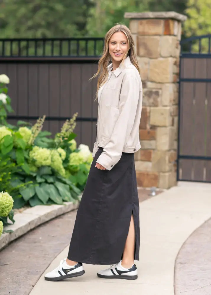 A woman wears a stella onyx black high waisted denim maxi skirt with a side slit paired with white and black sneakers