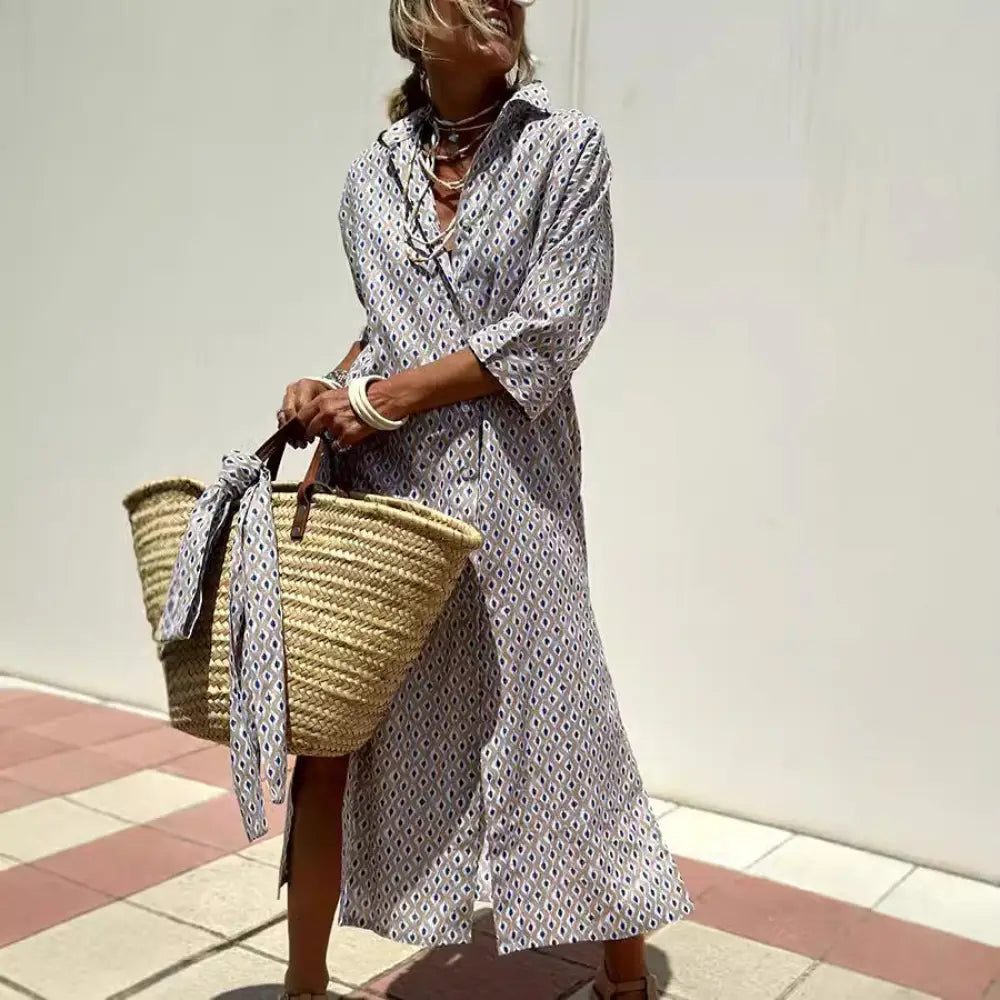 A woman wears a long light gray striped linen belted lapel shirt dress holding a woven straw tote bag