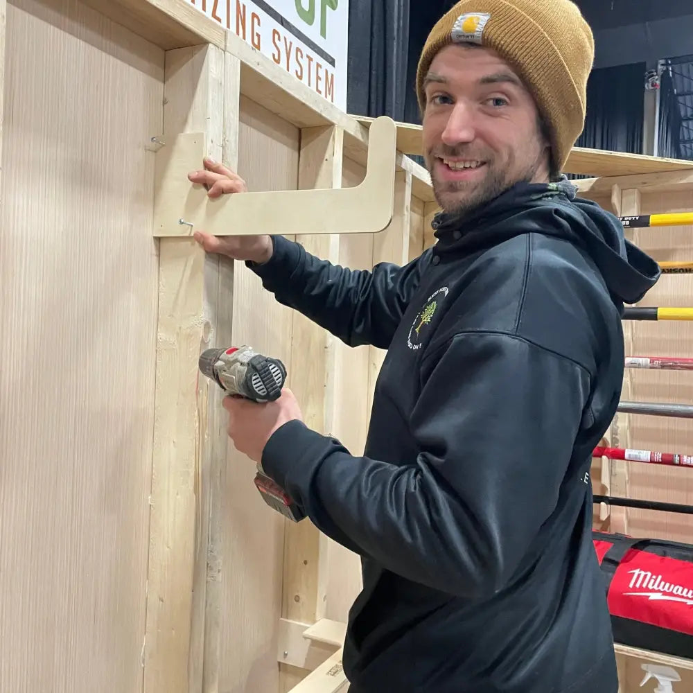 A man wearing a tan beanie and black hoodie drills wooden framing to install stud mounted wood and pipe shed storage hooks