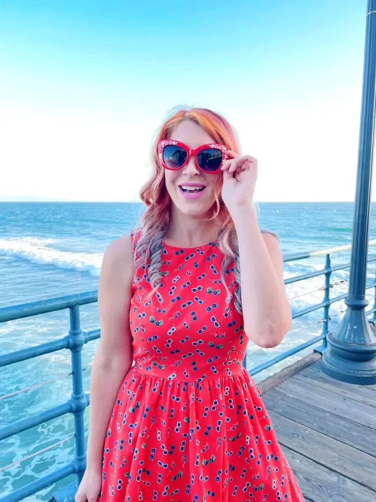 A woman wearing a vintage red floral sunglasses print cotton a line dress with bold red heart shaped sunglasses