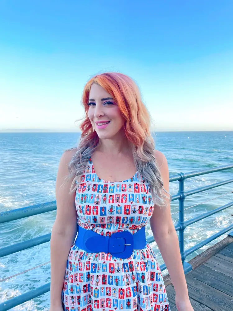 A woman wears a sleeveless sunny days vintage house print fit and flare dress with red white and blue postcard patterns