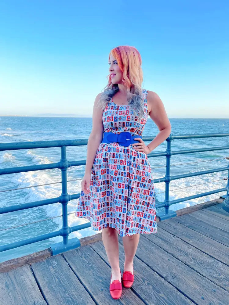 A woman wears a sleeveless knee-length sunny days vintage house print fit and flare dress with a royal blue belt