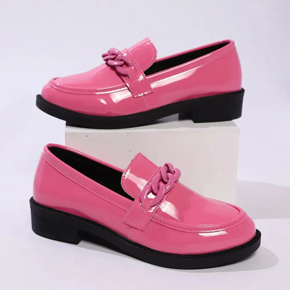 Bright pink patent leather loafers with black rubber sole and decorative chain-link strap, styled like olive green chunky chain loafers