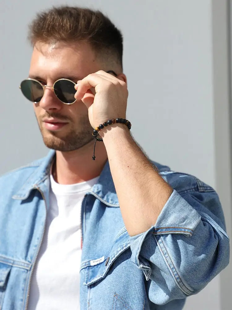 A man wearing round dark-lensed sunglasses with thin frames showcasing a Tulum tiger eye and black onyx beaded bracelet