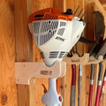 White and orange stihl hedge trimmer mounted on stud mounted wooden wall rack for string trimmers and tools