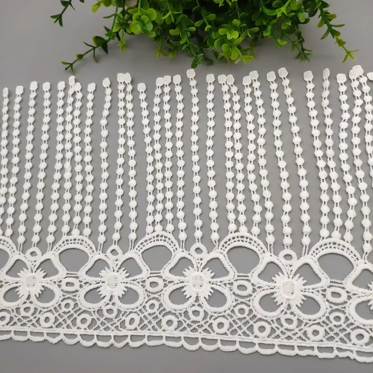 White beaded lace trim fabric featuring delicate beaded vertical strands and an ornate floral border in white lace