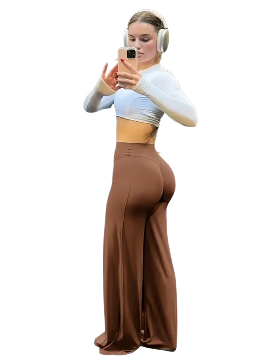 A woman wearing high waist wide leg brown fitness pants and a white long sleeved crop top standing in a gym