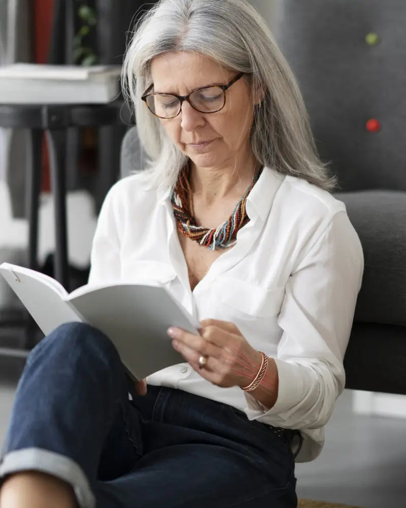 A woman with silver hair and glasses reads a book while wearing a copper magnetic therapy braided bangle bracelet