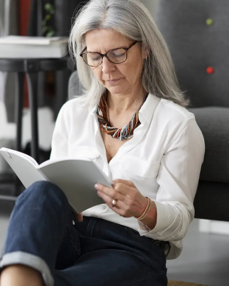 A woman with silver hair and glasses reads a book wearing a two-tone braided copper magnetic therapy bracelet