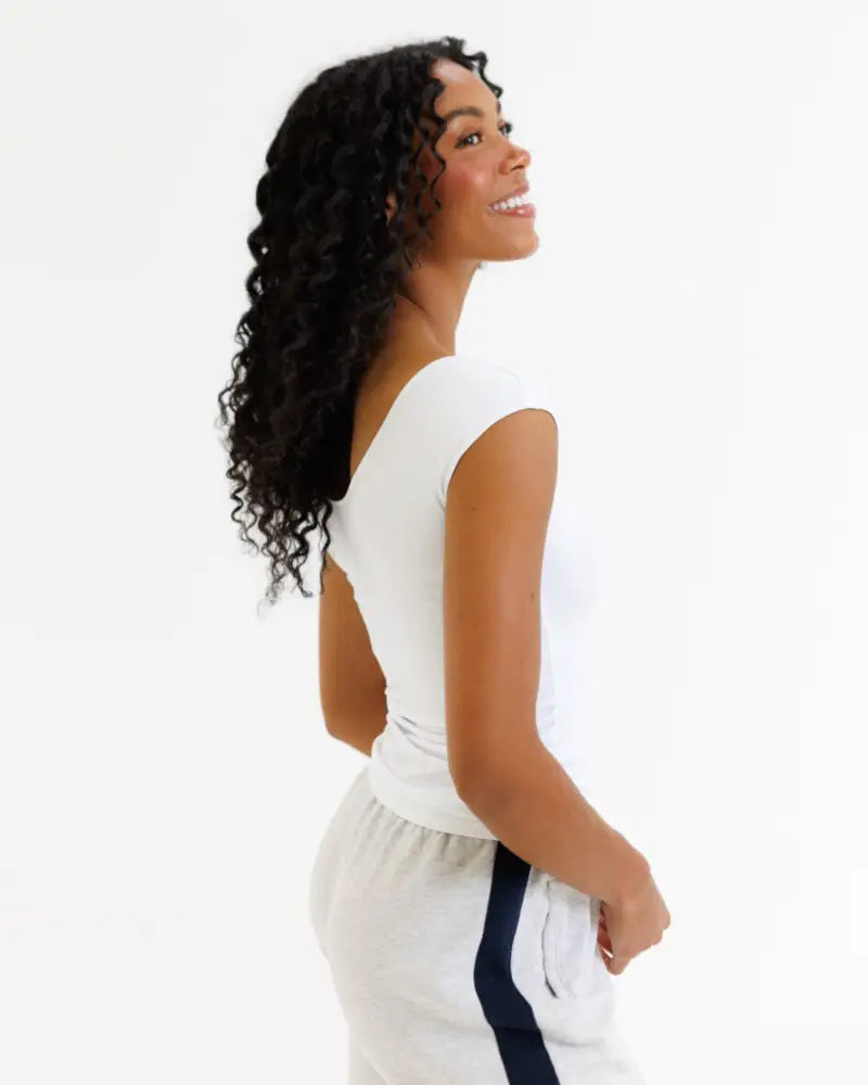 A woman wearing a white wonder tee cotton scoop neck cap sleeve t-shirt with a subtle back cutout and gray athletic pants