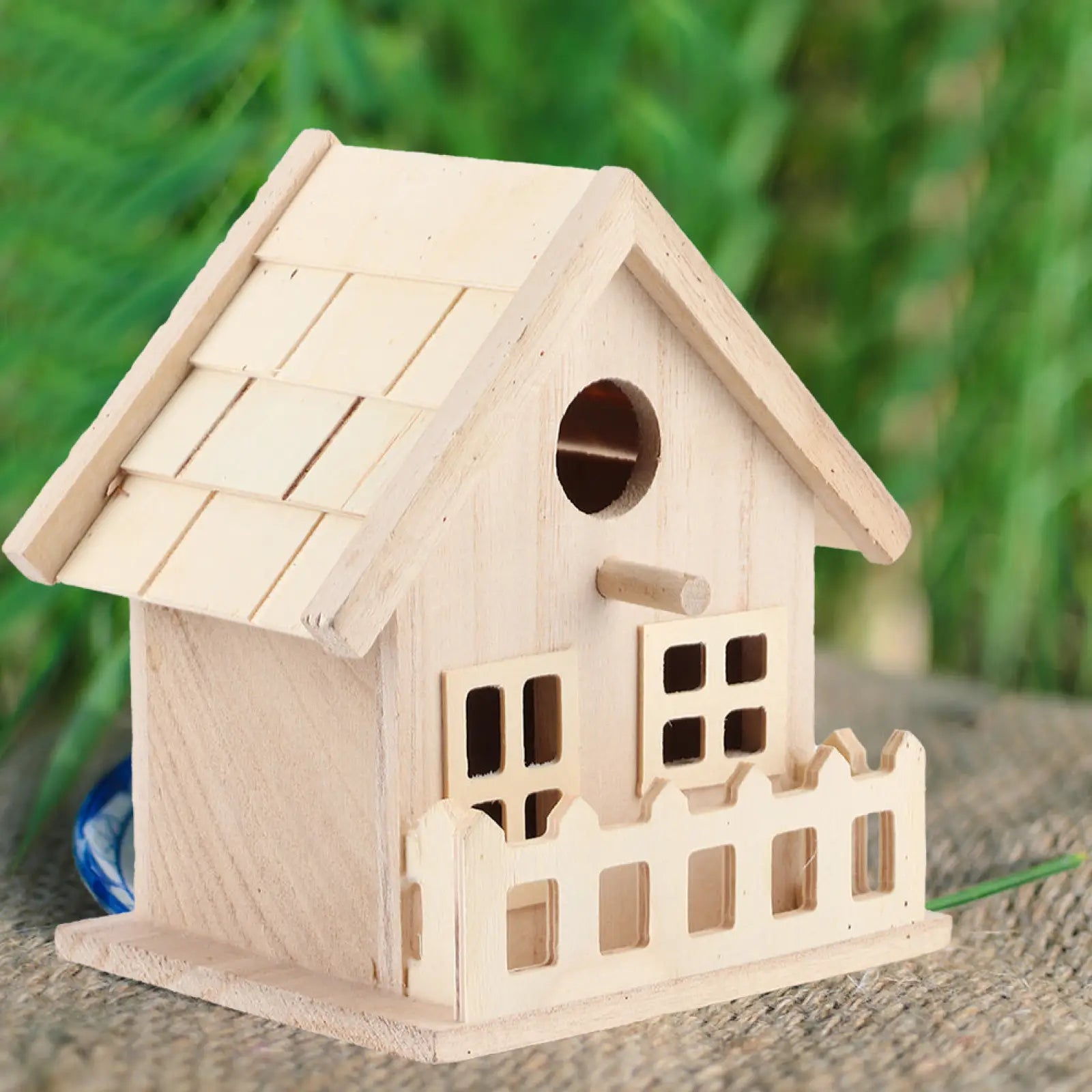 A light-colored wooden bird house with a slanted roof small windows and decorative fence base for parrots and small birds