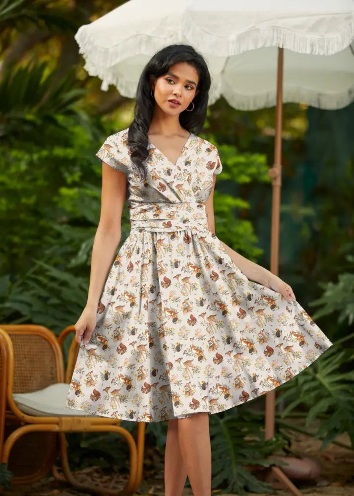 A woman wears a cream cotton woodland animals greta wrap dress with pockets featuring a v-neck and gathered waistband