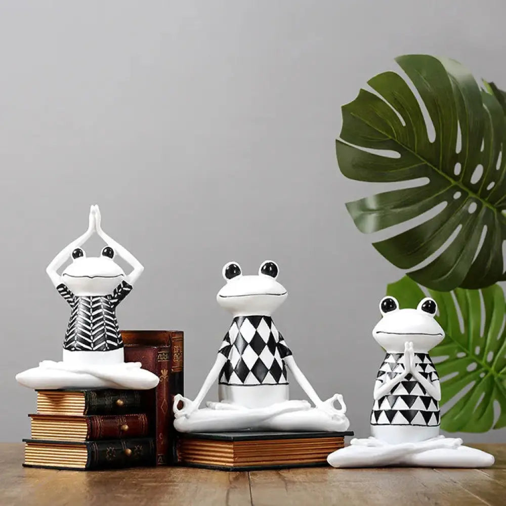 Three white resin frog figurines wearing black and white patterned shirts posed in different yoga positions for meditation