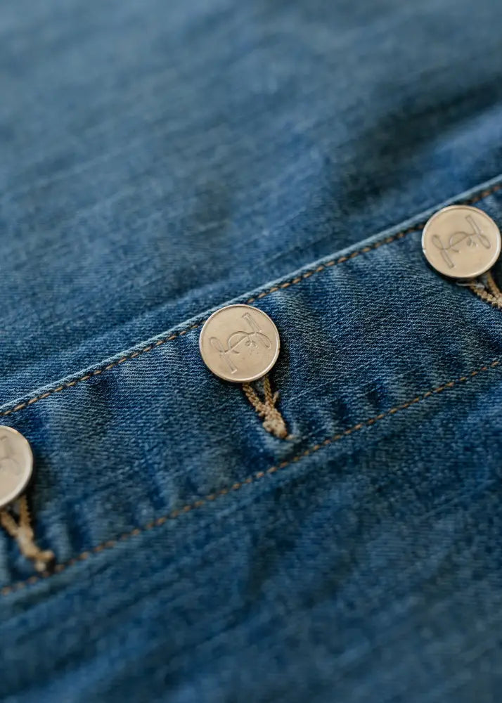 Silver coin-shaped buttons with engraved designs on blue denim fabric of zana front button denim midi skirt with kick pleat