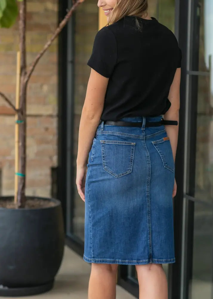 A mid-thigh length classic medium blue wash denim skirt with two back pockets and a black belt shown from the back
